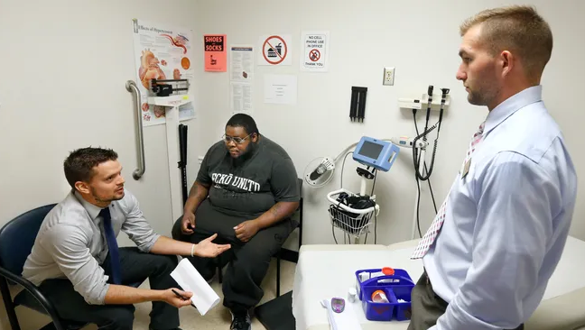 Two white doctors speak to a heavyset black man in a doctors office